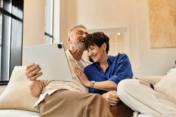 A happy couple sits closely on the couch, sharing laughter while looking at a tablet together. — Stock Photo