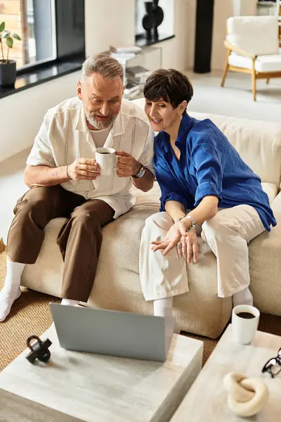 Middle aged couple shares laughter and warmth over coffee while engaged on their laptop. — Stock Photo