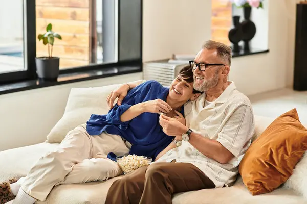 A happily married couple relaxes on their sofa while sharing laughter and popcorn at home. — Stock Photo