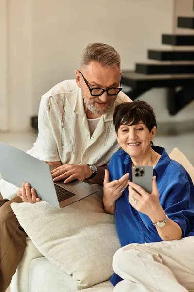 Coppia di mezza età si siede su un divano, sorridendo mentre si connettono tra loro utilizzando dispositivi. — Foto stock