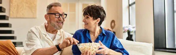Middle aged couple share laughter and popcorn on a cozy sofa at home in a joyful moment, banner — Stock Photo