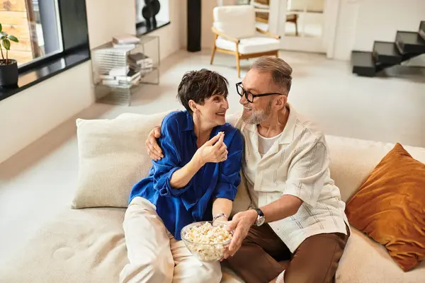 Middle-aged couple laughs and shares popcorn together in their cozy home. — Stock Photo