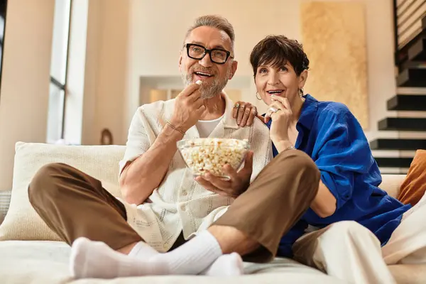 Middle aged couple enjoys popcorn together on a cozy couch, sharing laughter and warmth. — Stock Photo