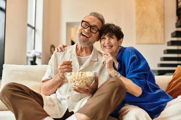 A married couple relaxes on their couch, sharing popcorn and laughter in their bright living room. — Stock Photo