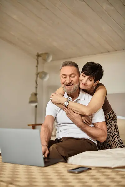 A happy middle aged couple embraces while exploring their laptop at home. — Stock Photo