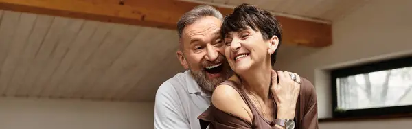 A joyful middle aged couple shares a warm embrace at home, smiling and laughing together, banner — Stock Photo