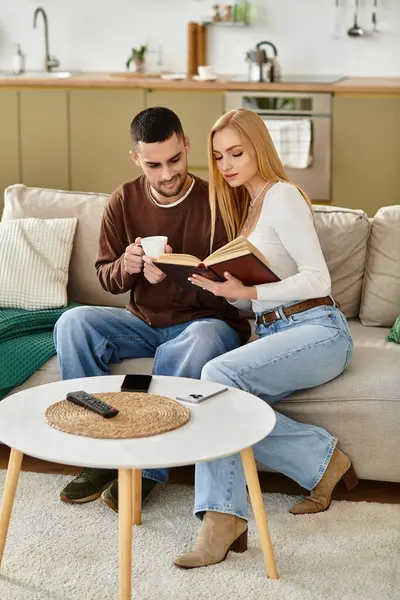A loving couple sits on their couch, sharing a cup of tea and reading a book in a warm home. — Stock Photo