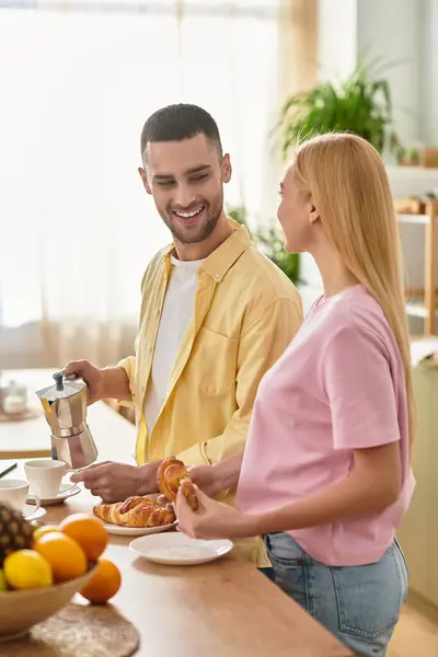 Bright light fills the kitchen as a man brews coffee while his partner enjoys a croissant, smiling. — Stock Photo