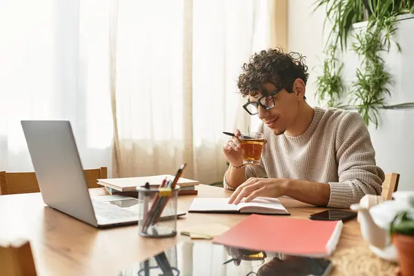 Handsome man takes a thoughtful sip from his cup as he writes in his notebook at home. — Stock Photo