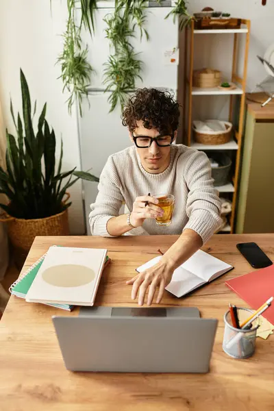A handsome young man sits at a wooden desk engaged in work, sipping a drink and taking notes. — Stock Photo