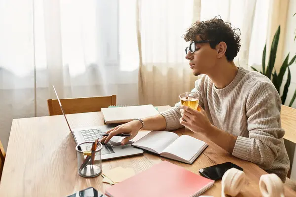 With a cup in hand, a fashionable young man focuses on his laptop surrounded by study materials. — Stock Photo