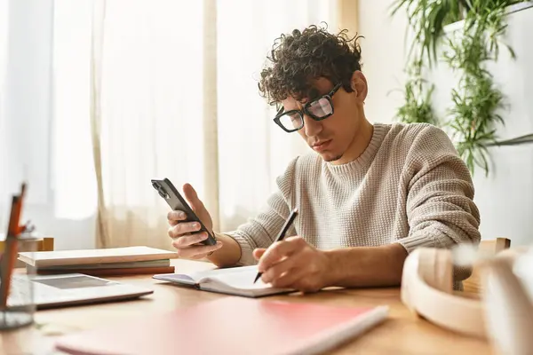 Beau jeune homme se concentre intensément sur l'écriture de notes tout en utilisant son smartphone à la maison. — Photo de stock