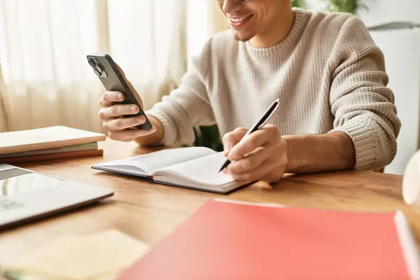 A stylish young man smiles while jotting notes in a cozy, book filled space with a smartphone. — Stock Photo