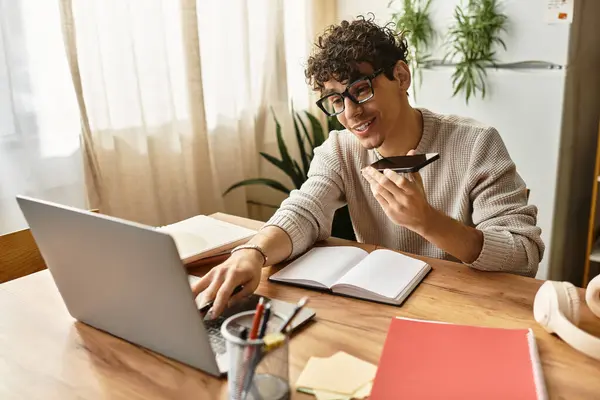 Un bel ragazzo parla in uno smartphone mentre si concentra su un computer portatile su una scrivania di legno. — Foto stock