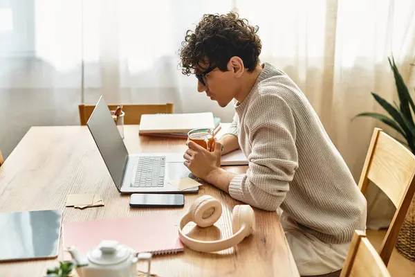 Elegante joven macho se centra en su portátil, rodeado de cuadernos y una bebida refrescante. - foto de stock