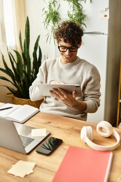 Young man with curly hair, wearing glasses, happily working on tablet in his modern workspace. — Stock Photo