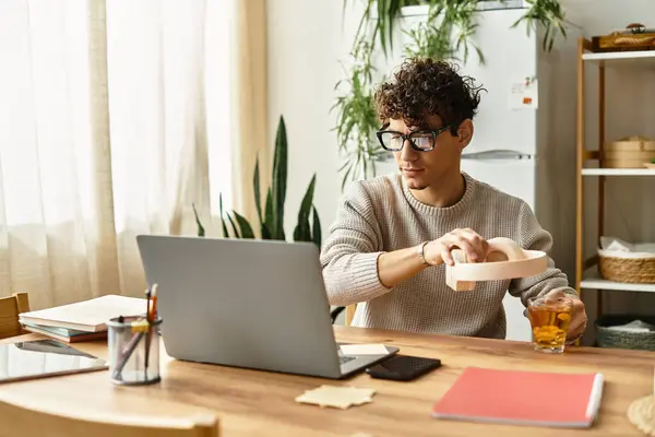 Bello uomo in un maglione accogliente prepara il tè mentre lavora con attenzione al suo computer portatile. — Foto stock