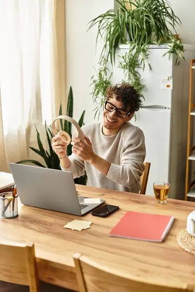 Cheerful man with curly hair smiles while holding headphones, working at a warm wooden table. — Stock Photo