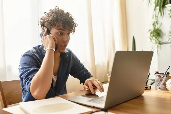 Giovane uomo in abbigliamento casual chatta al telefono mentre si concentra sul suo computer portatile in una zona luminosa. — Foto stock
