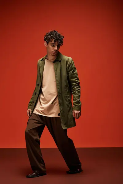 A handsome man with curly hair showcases a modern outfit while standing against a bold red backdrop. — Stock Photo