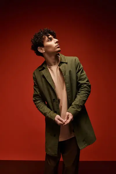 Giovane con i capelli ricci indossa un cappotto alla moda, guardando pensieroso mentre in piedi contro il rosso. — Foto stock