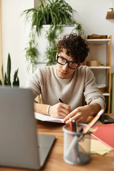 Jeune homme aux cheveux bouclés écrit des notes tout en se concentrant sur les études dans un espace de travail confortable. — Photo de stock