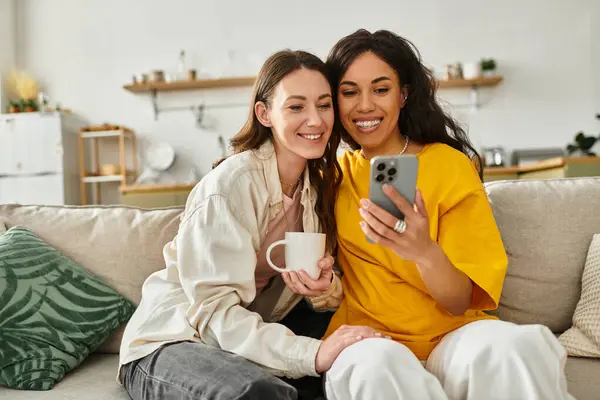 Two women enjoy each others company, looking at a phone together while relaxing at home. — Stock Photo