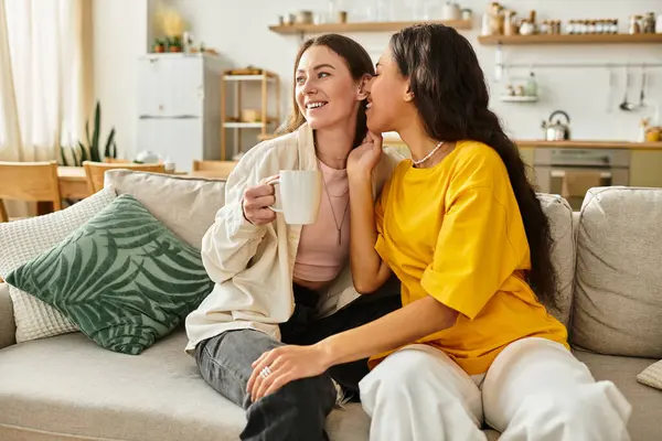 Two women sit close on the sofa, laughing and sharing intimate whispers while enjoying tea. — Stock Photo