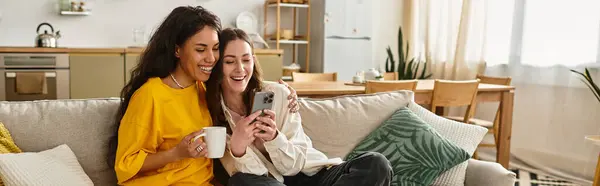 Due donne si abbracciano amorevolmente sul divano, ridendo mentre usano uno smartphone. — Stock Photo