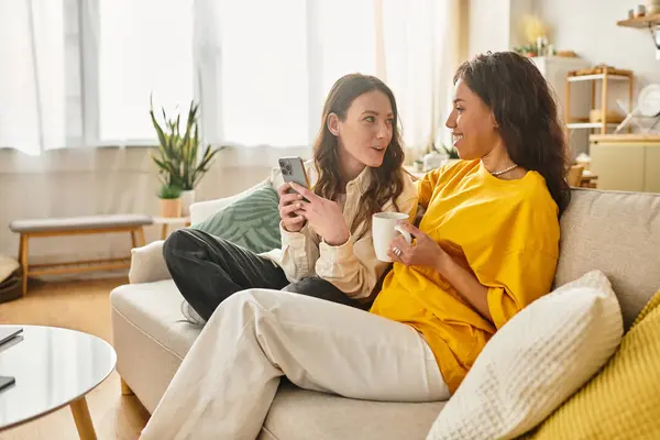Two women relax on a couch, sharing a warm drink and joyous conversation in their cozy living space. — Stock Photo