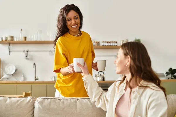 Duas mulheres sorriem calorosamente enquanto trocam xícaras de café em seu espaço de vida convidativo. — Fotografia de Stock
