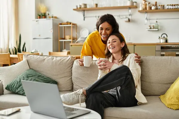 Due donne sedute vicino a un divano, ridendo e guardando uno spettacolo con bevande calde in mano. — Foto stock