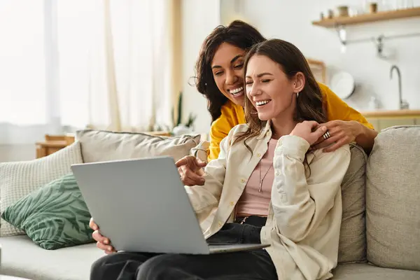Zwei Frauen lächeln und lachen zu Hause zusammen, während sie sich einen Laptop teilen, umgeben von Wärme. — Stockfoto