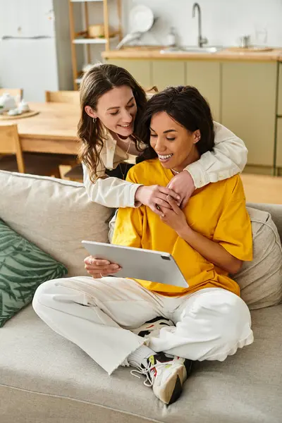 Two women share laughter and warmth as they look at a tablet, embracing affectionately at home. — Stock Photo