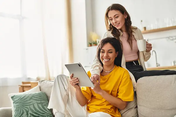 Two women cuddle on a sofa, joyfully exploring content on a tablet in their cozy living space. — Stock Photo