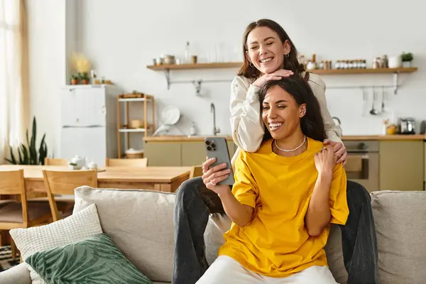 Two women share a joyful moment at home, cuddling on the couch and looking at a phone together. — Stock Photo