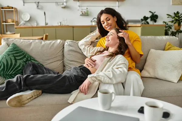 Two loving women share tender laughter while relaxing on their couch at home together. — Stock Photo