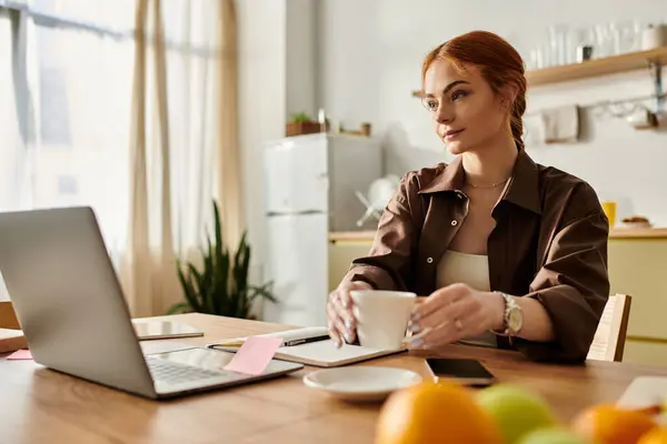 A red haired woman sits at a wooden table, holding a coffee cup and focusing on her laptop. — Stock Photo