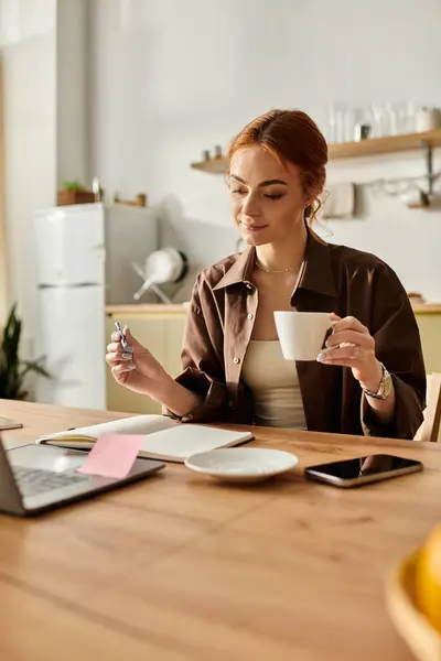 Femme aux cheveux roux saisissants tient une tasse de café, engagé dans l'écriture de notes à une table en bois. — Photo de stock