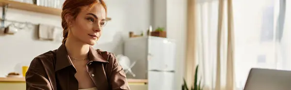 Красивая женщина с рыжими волосами фокусируется на ноутбуке, сидя в теплой кухне. — стоковое фото