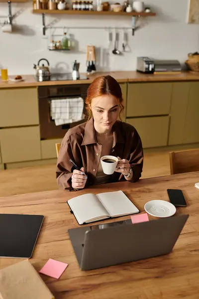 Red haired woman engaged in writing notes while sipping coffee in a warm and welcoming kitchen. — Stock Photo