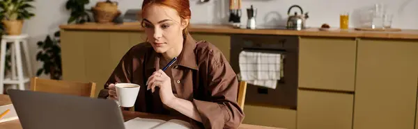 Сосредоточенная женщина с рыжими волосами пьет кофе, внимательно изучая ноутбук на теплой кухне. — стоковое фото