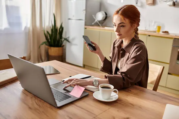 A red haired woman sits at a wooden table, focused on her laptop and phone with coffee. — Stock Photo