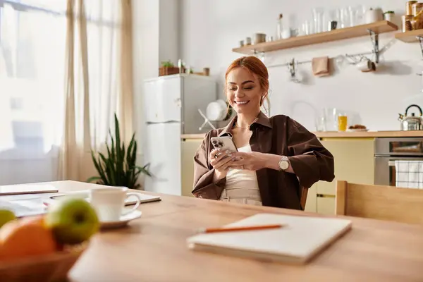A cheerful woman with red hair checks her phone at a wooden table in a lovely kitchen. — Stock Photo
