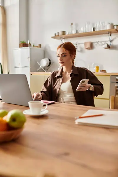 A woman with red hair works intently on her laptop at a sunny kitchen table. — Stock Photo