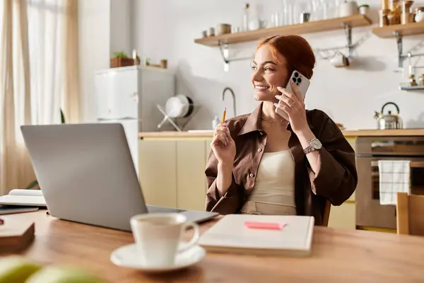 Una donna allegra con i capelli rossi parla al telefono mentre è seduta a un tavolo da cucina. — Foto stock