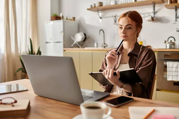 Red haired woman thoughtfully writes in a notebook while seated at a rustic kitchen table. — Stock Photo