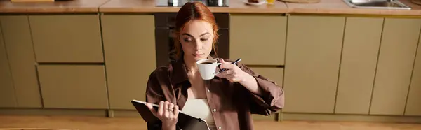 A beautiful red haired woman enjoys her coffee and writes in a notebook in a warm kitchen. — Stock Photo