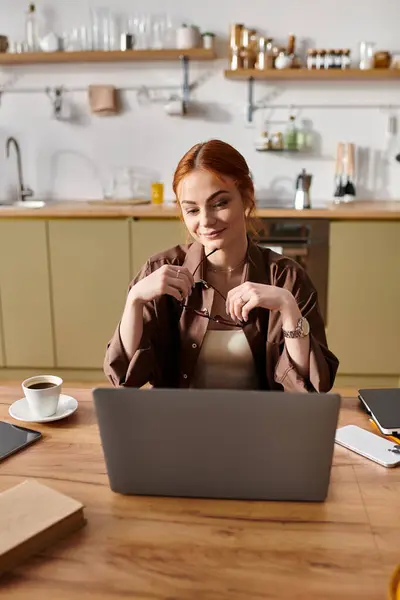 Beautiful red haired woman smiles while working on her laptop in a cozy kitchen environment. — Stock Photo