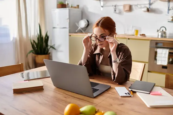 Una hermosa mujer pelirroja ajusta sus gafas mientras se concentra en su computadora portátil en casa. - foto de stock